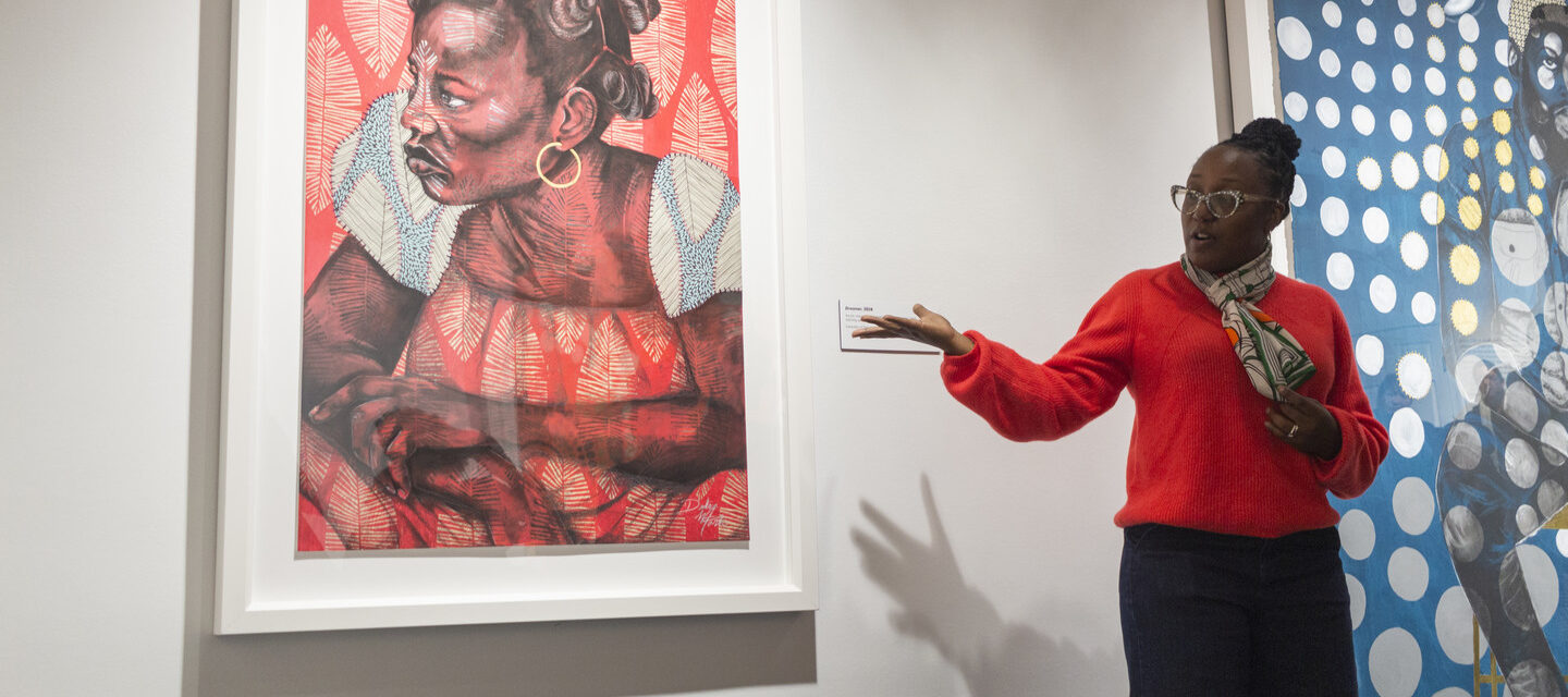 The artist, a woman with dark skin tone, speaks in front of her artwork, gesturing to it, a large colorful print of a woman with a pattern overlay across the paper. The artist wears a bright right sweater, a multicolored scarf, glasses, and gold hoops.