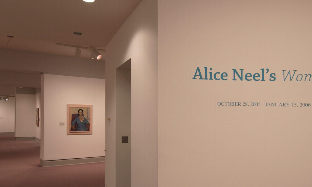 An installation view of a gallery space shows several paintings hanging on a white wall. On another wall, it says in big blue letters: 