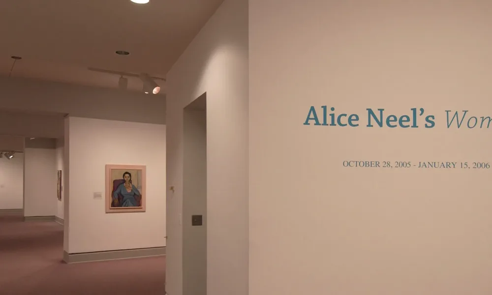 An installation view of a gallery space shows several paintings hanging on a white wall. On another wall, it says in big blue letters: "Alice Neel's Women".