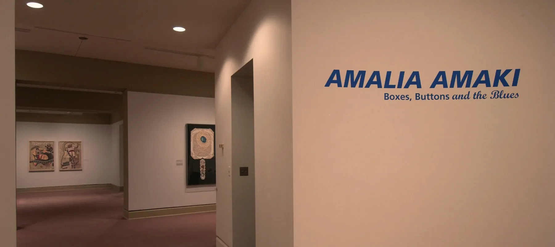 Installation view of a gallery space. On a white wall, big letters in blue say: "Amalia Amaki: Boxes, Buttons, and the Blues". On the white walls behind this wall, several art pieces are hanging.