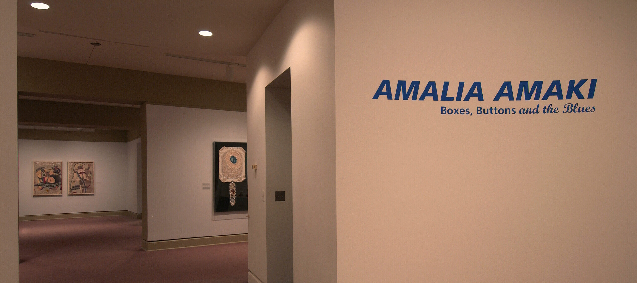 Installation view of a gallery space. On a white wall, big letters in blue say: 