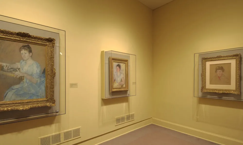 Installation view of a gallery space. One large and two smaller paintings and drawings are hanging on a yellow wall behind glass. The paintings depict women and are painted in an Impressionist style, with smooth brushstrokes and in light colors.