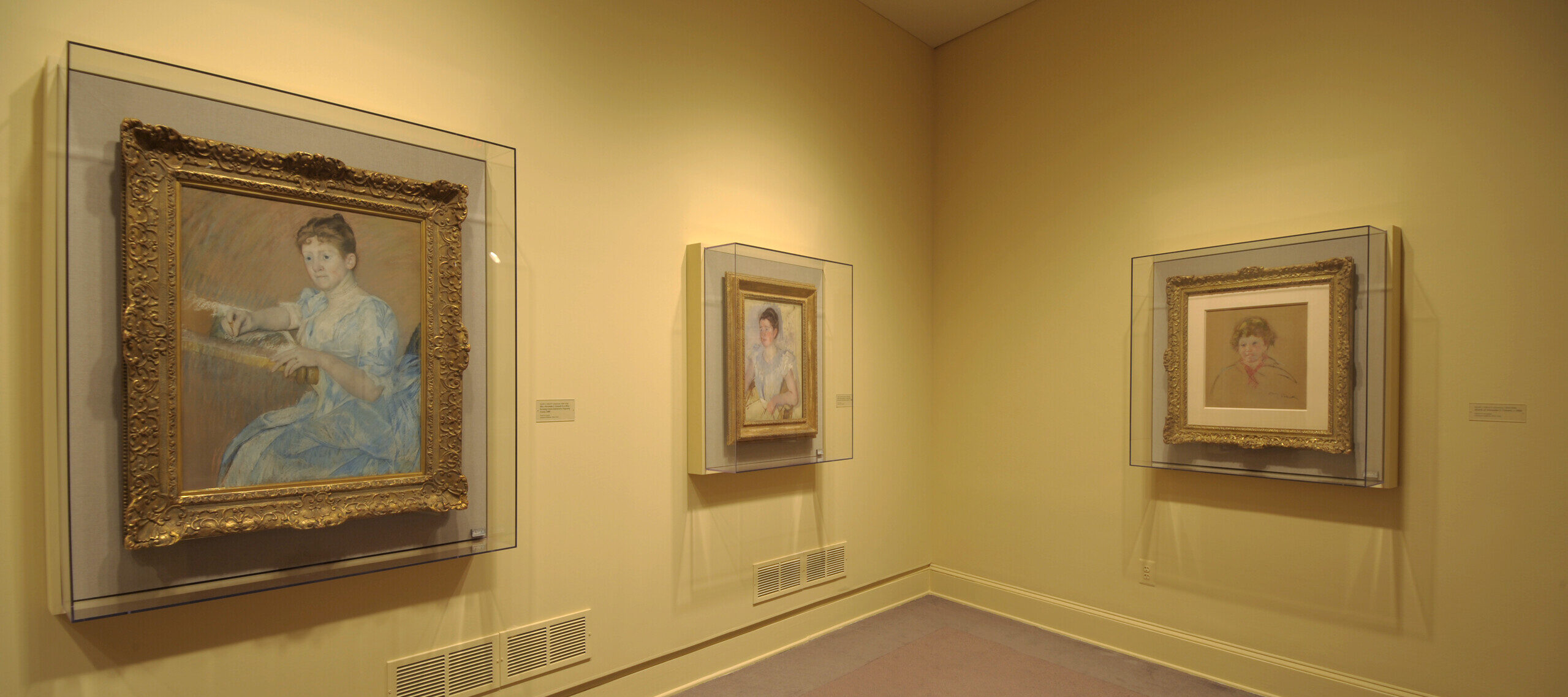 Installation view of a gallery space. One large and two smaller paintings and drawings are hanging on a yellow wall behind glass. The paintings depict women and are painted in an Impressionist style, with smooth brushstrokes and in light colors.