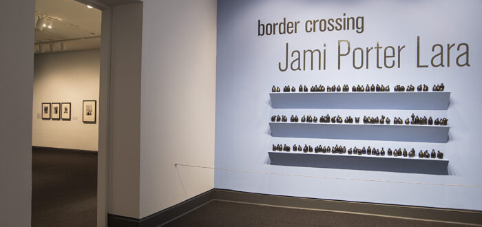 Installation view of a gallery space. On a bright blue wall, it says in big black letters "Border crossing" and "Jami Porter Lara". Underneath are three exposed shelves covered in small black bottle sculptures.
