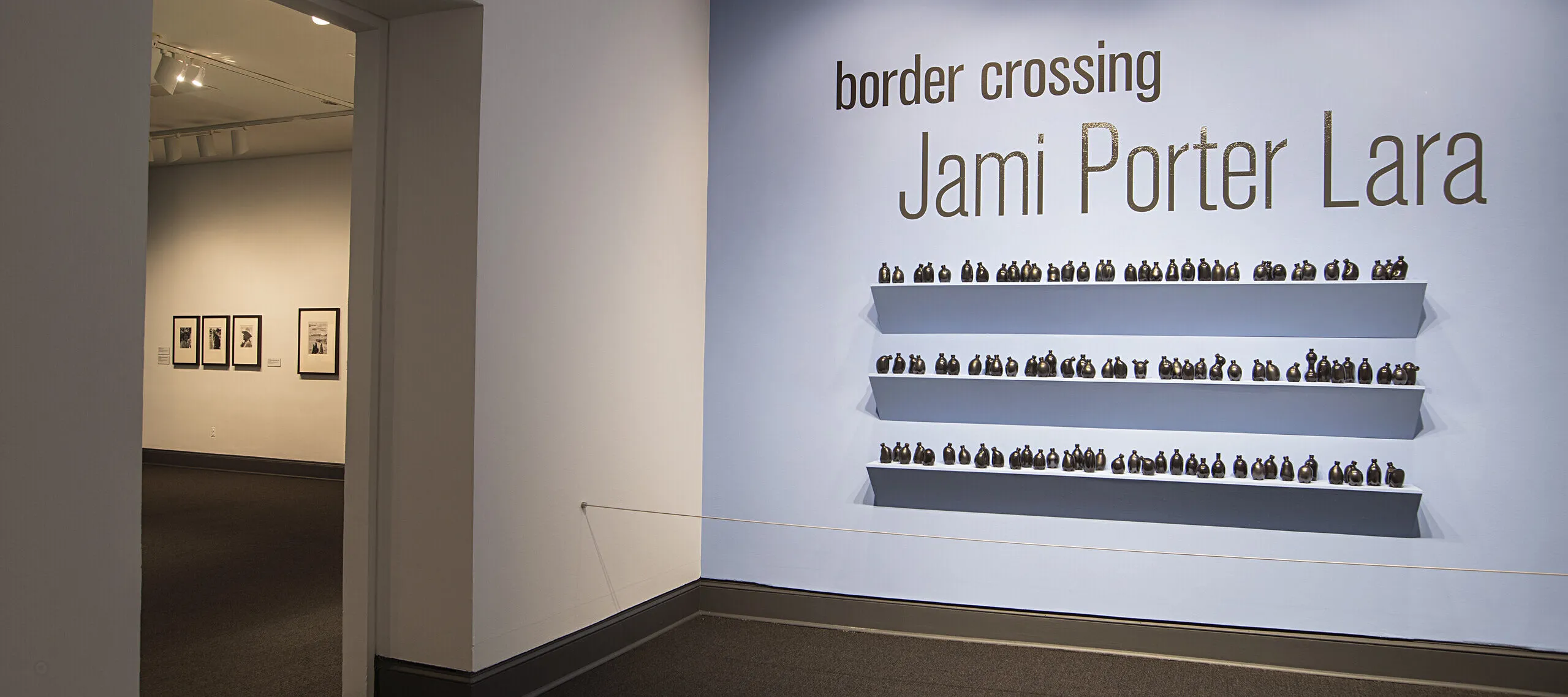 Installation view of a gallery space. On a bright blue wall, it says in big black letters "Border crossing" and "Jami Porter Lara". Underneath are three exposed shelves covered in small black bottle sculptures.