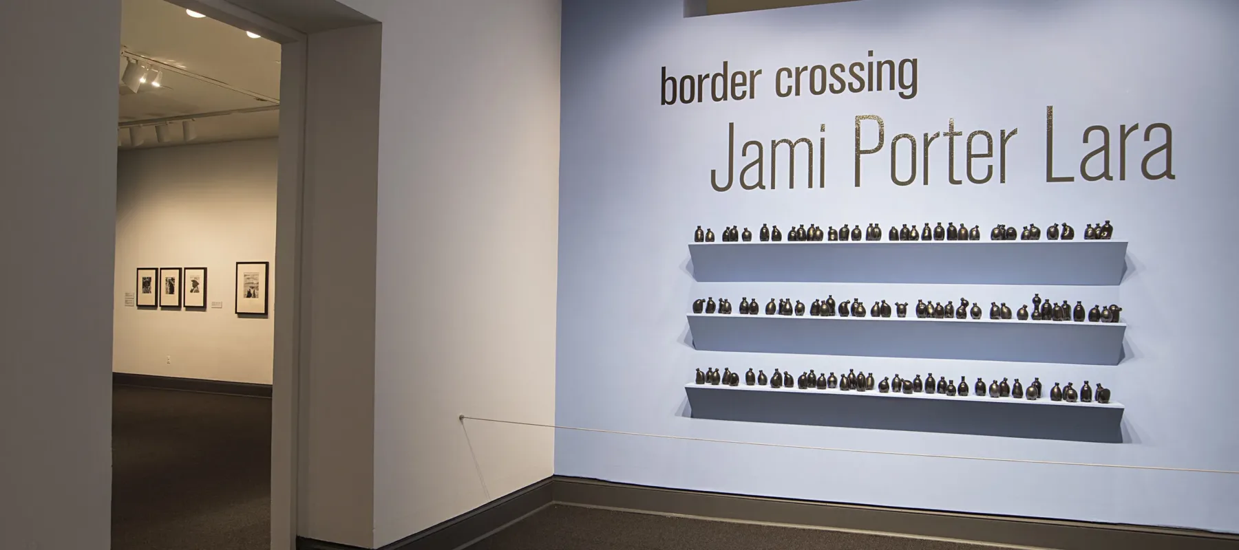 Installation view of a gallery space. On a bright blue wall, it says in big black letters "Border crossing" and "Jami Porter Lara". Underneath are three exposed shelves covered in small black bottle sculptures.