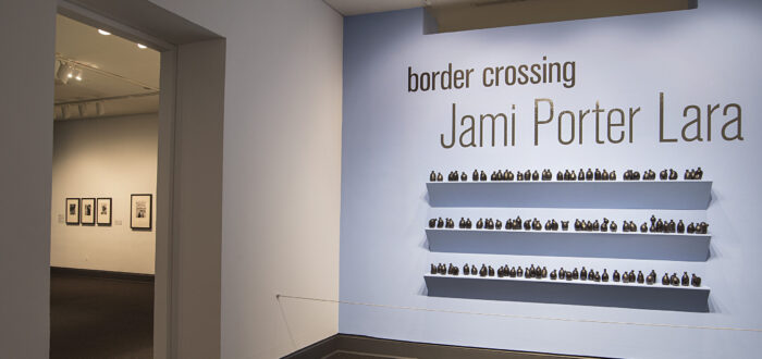 Installation view of a gallery space. On a bright blue wall, it says in big black letters "Border crossing" and "Jami Porter Lara". Underneath are three exposed shelves covered in small black bottle sculptures.