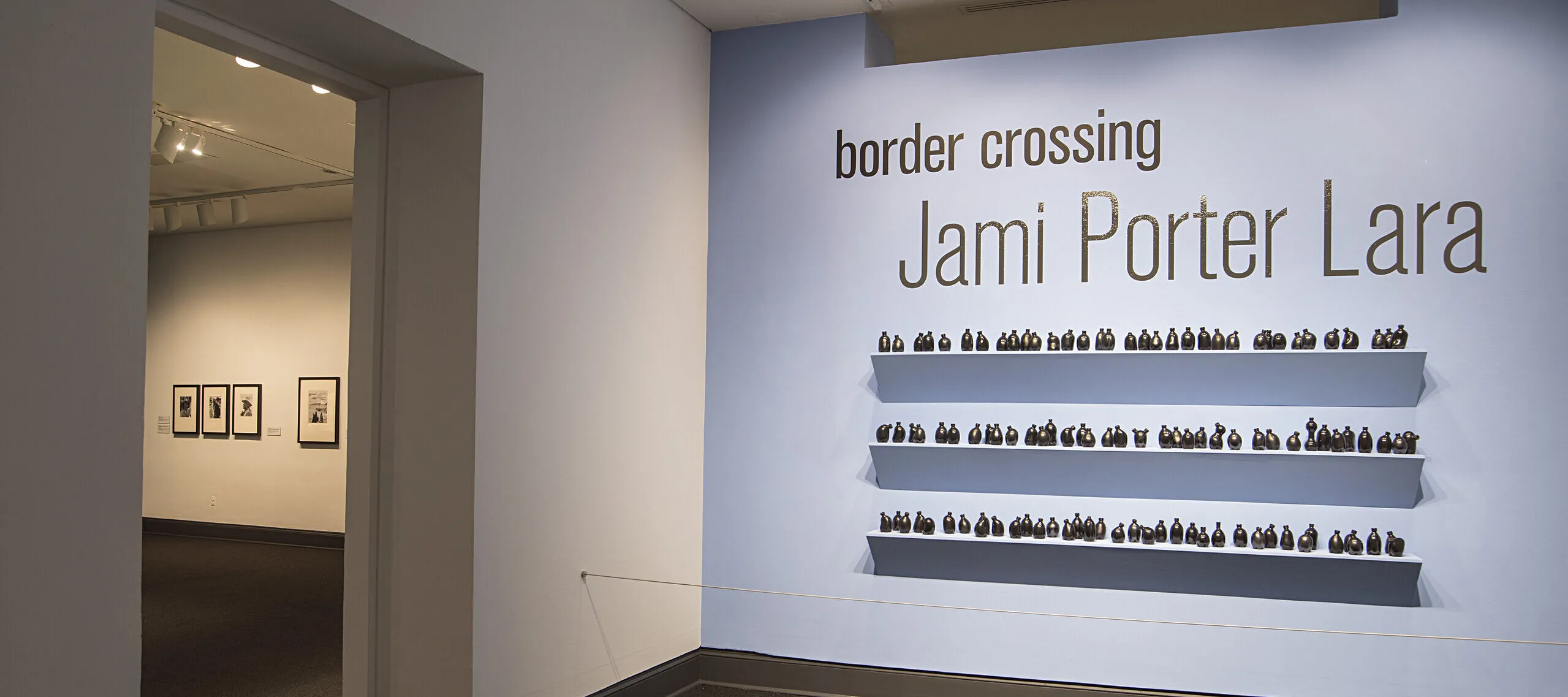 Installation view of a gallery space. On a bright blue wall, it says in big black letters "Border crossing" and "Jami Porter Lara". Underneath are three exposed shelves covered in small black bottle sculptures.