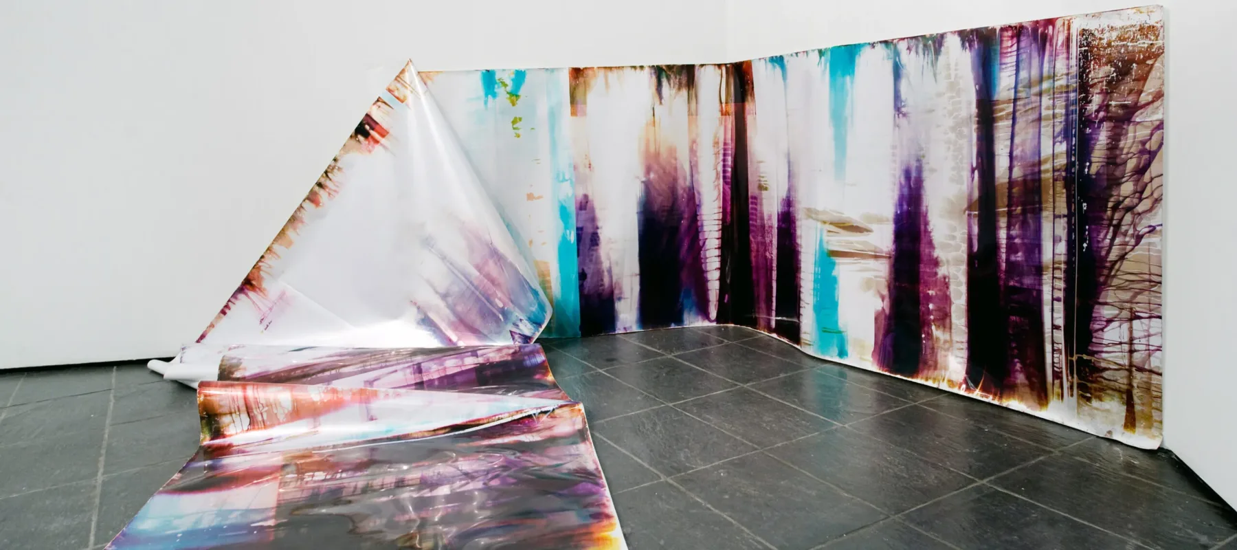 In an art gallery with white walls, a long, snake-like piece of metallic paper printed with splotches of black, blue, purple, and orange in an abstract pattern is partly affixed to the wall. At one point the paper falls from the wall and cascades onto the floor and further into the room.