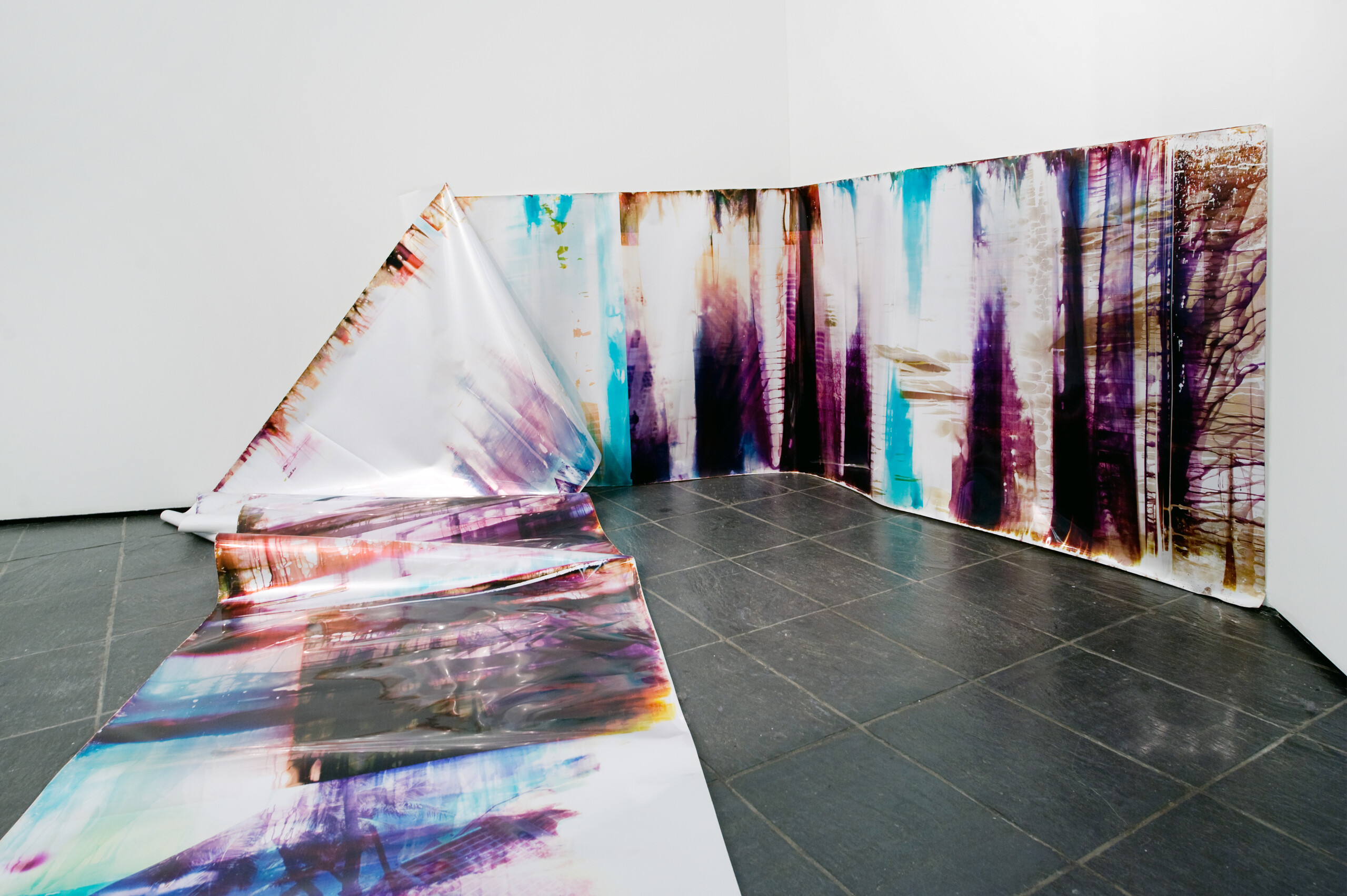 In an art gallery with white walls, a long, snake-like piece of metallic paper printed with splotches of black, blue, purple, and orange in an abstract pattern is partly affixed to the wall. At one point the paper falls from the wall and cascades onto the floor and further into the room.