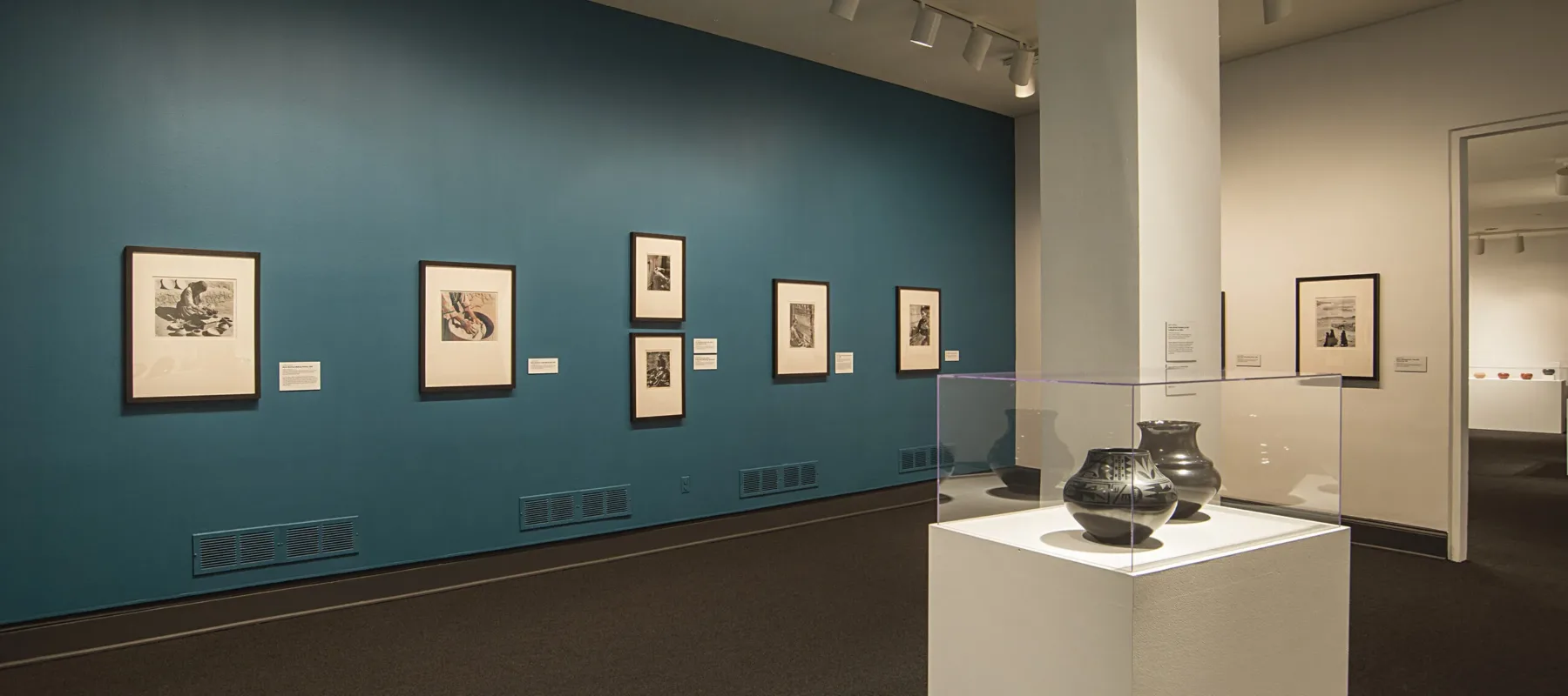 Installation view of a gallery space featuring black and white photographs hanging on a blue wall and black ceramic or clay vessels placed on pedestals in a glass box.