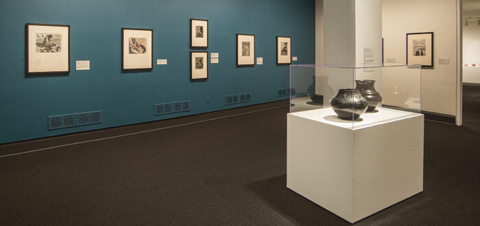 Installation view of a gallery space featuring black and white photographs hanging on a blue wall and black ceramic or clay vessels placed on pedestals in a glass box.