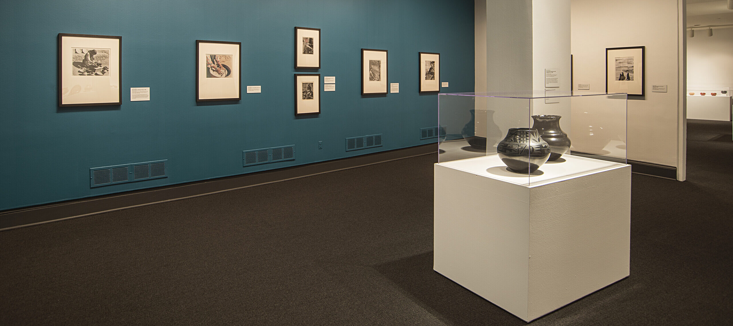 Installation view of a gallery space featuring black and white photographs hanging on a blue wall and black ceramic or clay vessels placed on pedestals in a glass box.