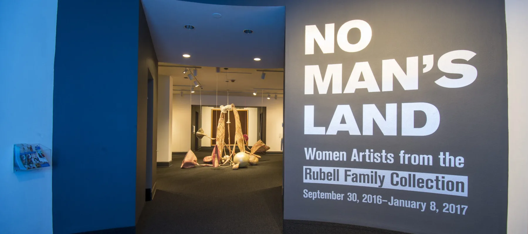 View of a gallery space. On a black wall, it says in white, bold letters: "NO MAN’S LAND: Women Artists from the Rubell Family Collection". There is a textile sculpture hanging from the ceiling in the room behind the wall.