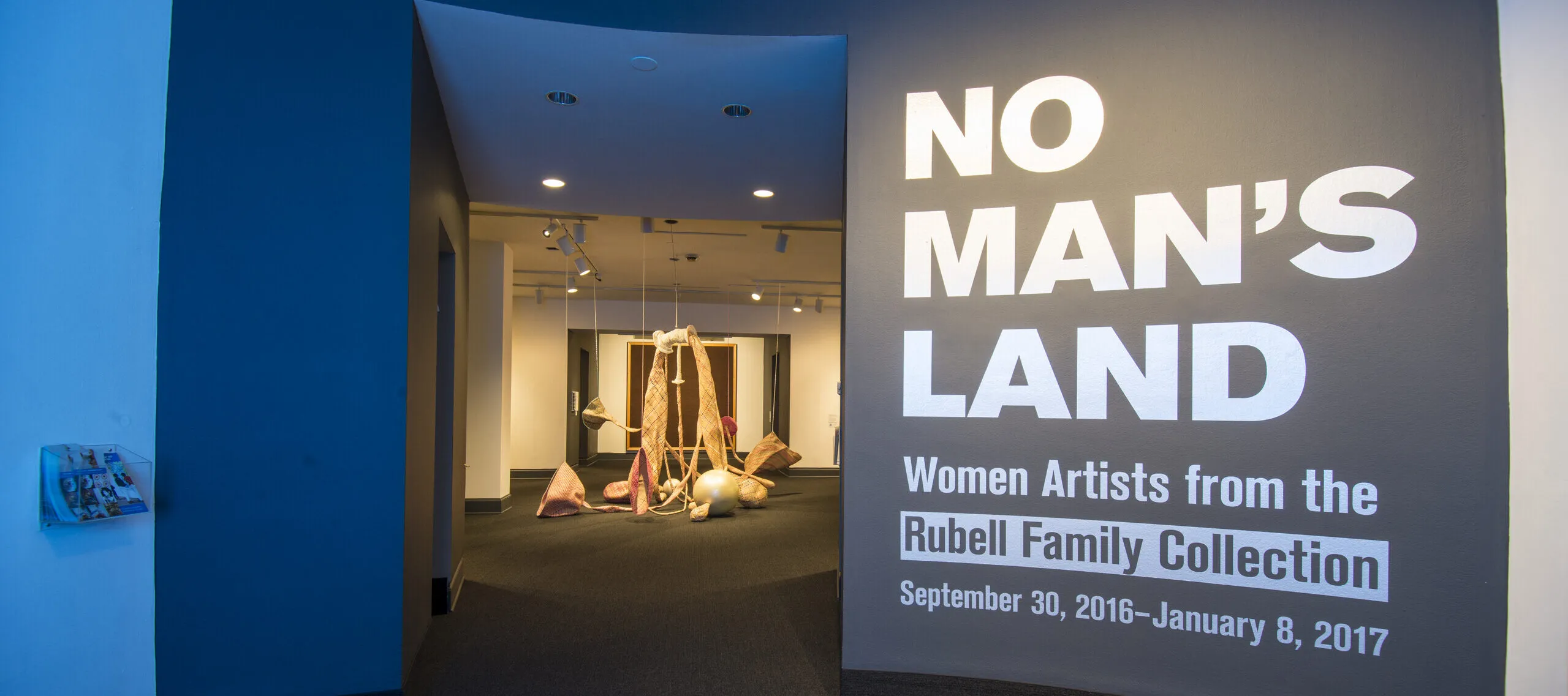 View of a gallery space. On a black wall, it says in white, bold letters: "NO MAN’S LAND: Women Artists from the Rubell Family Collection". There is a textile sculpture hanging from the ceiling in the room behind the wall.