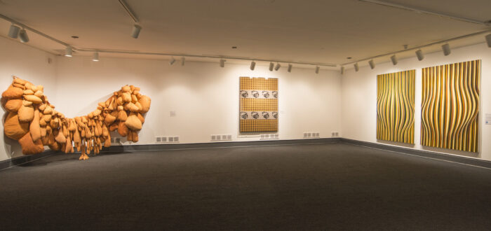 An installation view of a gallery space with white walls and a gray floor. There are several art pieces hanging on the walls. On the left, a huge textile piece made of several bags in an orange fabric takes up a big part of the room. Another art piece shows skulls on an orange checkered background. On the right wall is an abstract art piece made of several orange, white, and black stripes that causes an optical illusion.