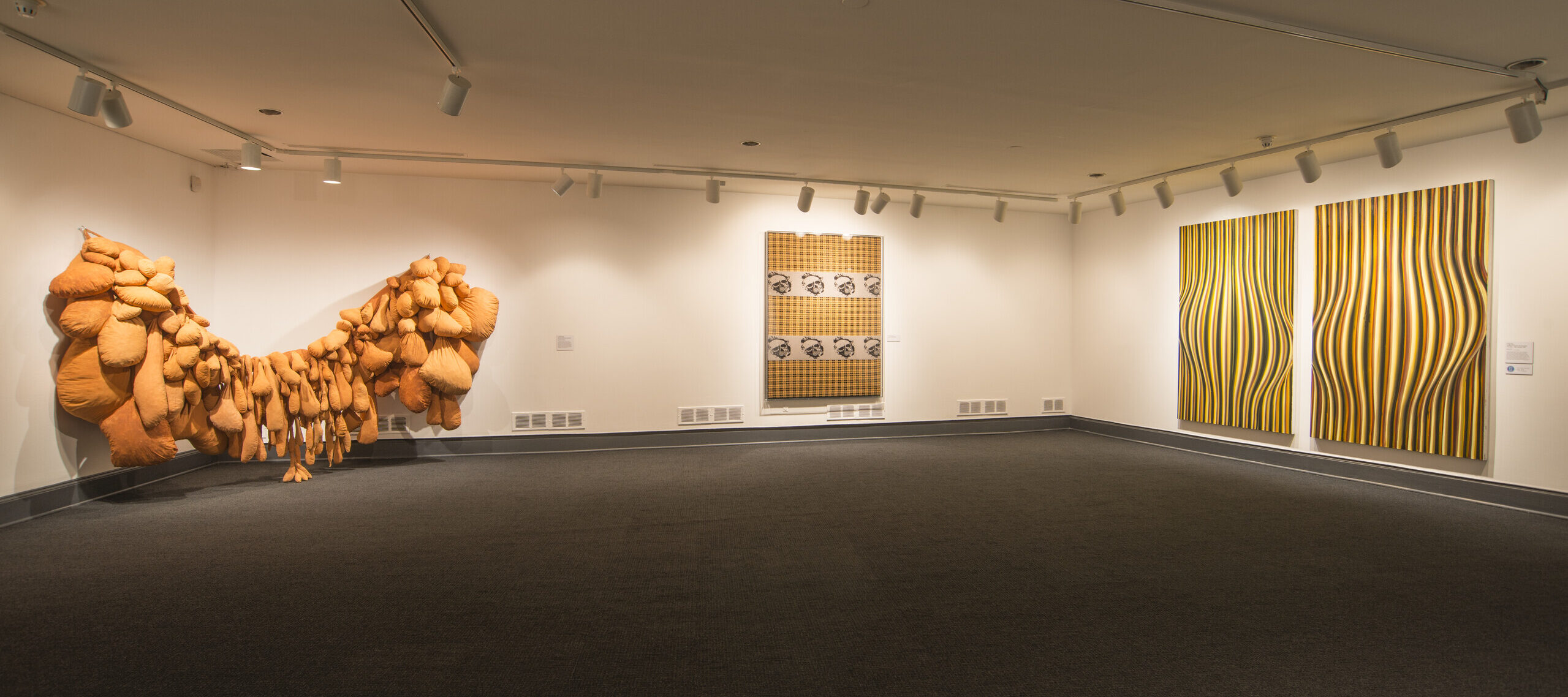 An installation view of a gallery space with white walls and a gray floor. There are several art pieces hanging on the walls. On the left, a huge textile piece made of several bags in an orange fabric takes up a big part of the room. Another art piece shows skulls on an orange checkered background. On the right wall is an abstract art piece made of several orange, white, and black stripes that causes an optical illusion.