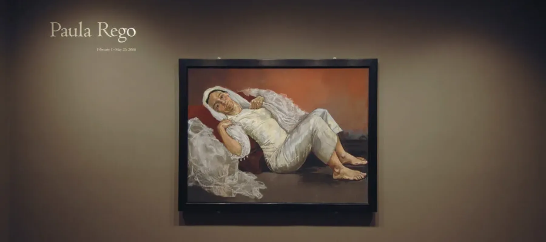 A view of a gallery space shows a large painting on a beige wall and in big letters next to it, it says "Paula Rego". The painting shows a woman in white clothing leaning back while sitting on the floor. She is wearing a wedding dress and a veil, and next to her shoulders, draped pieces of fabric are placed in a way that makes it look she has angel wings.