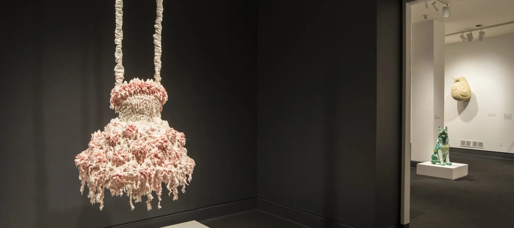 Installation view of a sculpture hanging from the ceiling in a gallery space. The sculpture consists of myriad layers of melted pink and white wax that encrust and obscure the metal armature for this abstract sculpture, which hangs from satin-wrapped chains. Its color and shape, as well as the bumpy, lacy texture, evoke a frilly tutu, lavishly frosted wedding cake, or coral accretions.