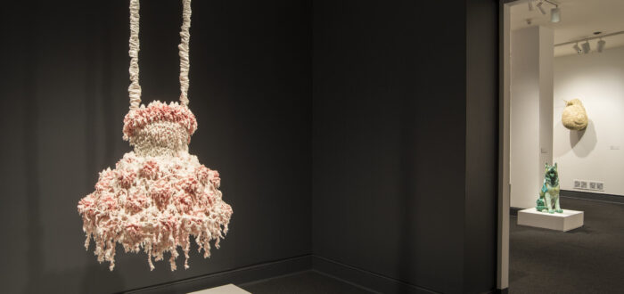 Installation view of a sculpture hanging from the ceiling in a gallery space. The sculpture consists of myriad layers of melted pink and white wax that encrust and obscure the metal armature for this abstract sculpture, which hangs from satin-wrapped chains. Its color and shape, as well as the bumpy, lacy texture, evoke a frilly tutu, lavishly frosted wedding cake, or coral accretions.