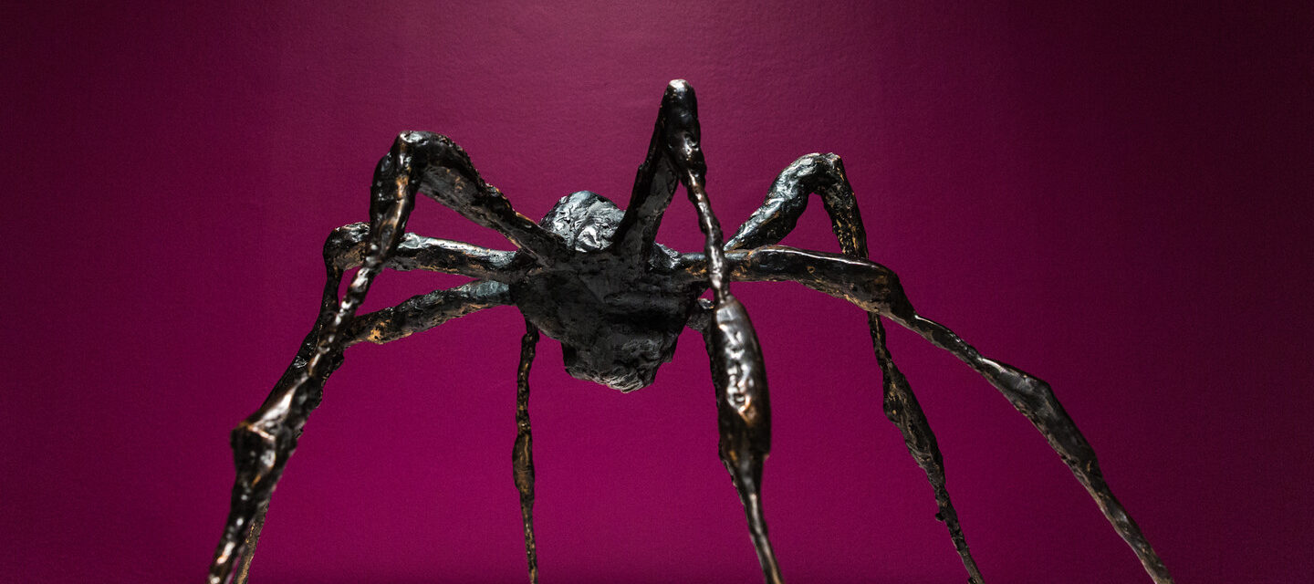Bronze sculpture of a spider on a white platform against a magenta background.