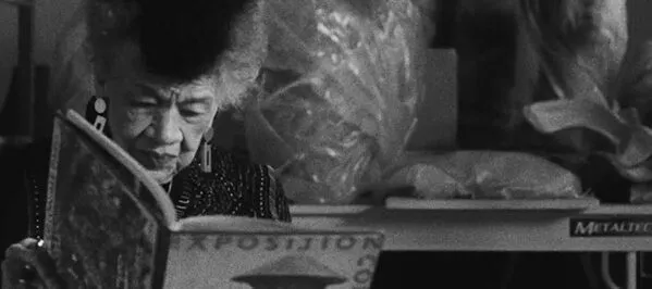 A black and white photograph of a medium-skinned woman with short grey hair that has a dark brown streak down the middle. She is wearing a dark beaded jacket and sits in front of a table filled with sculptures, reading a large book.