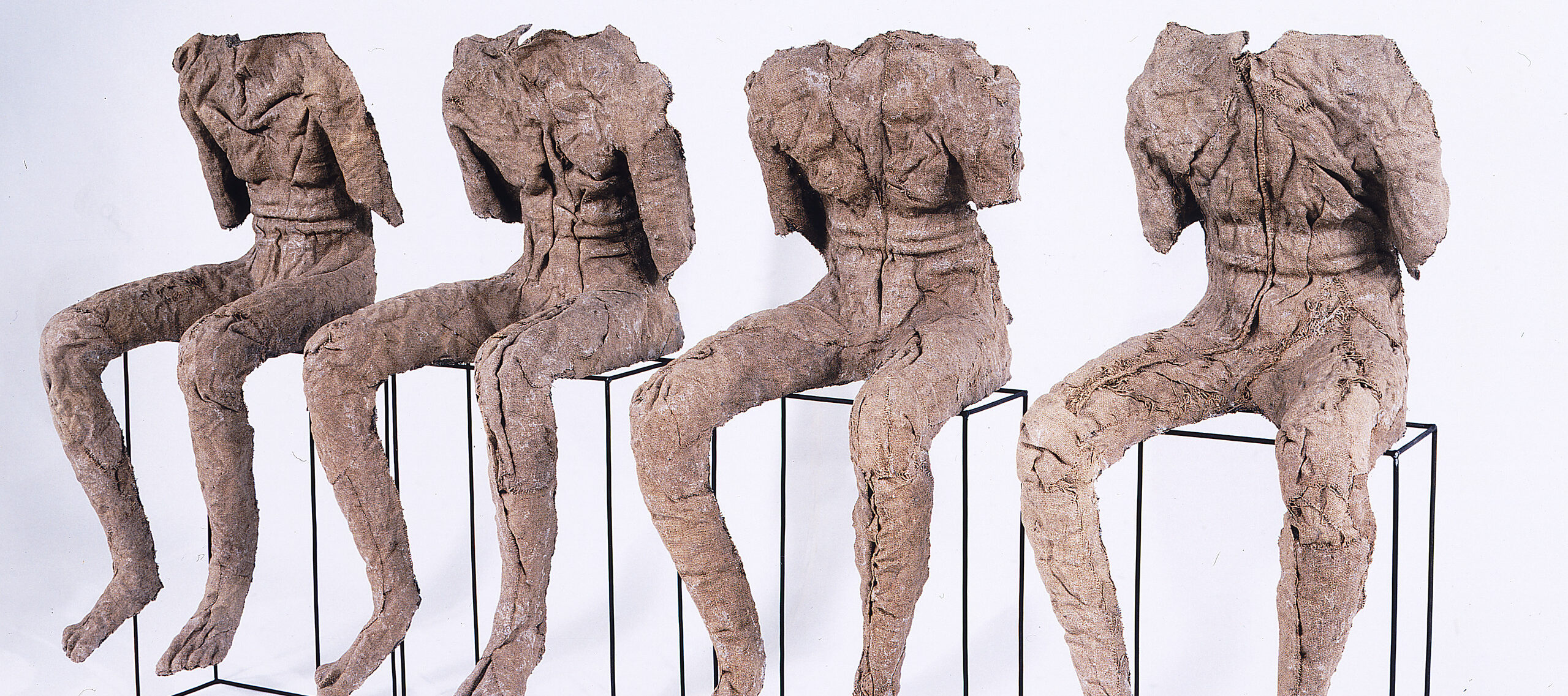 Four androgynous figures rendered in stiffened, brown burlap sit atop vertical, rectangular metal frames. They lack necks, heads, lower arms, and clothing, and their upper torsos slump slightly forward. The burlap’s color and bumpy texture evoke bark and mummy wrappings.