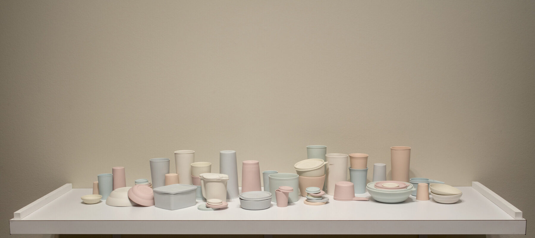 A collection of pastel-colored porcelain cups, bowls, containers, and lids that resemble plastic food containers. Some are stacked within and on top of each other. Some containers have lids firmly shut while on others the lids teeter off the edge of container rims.