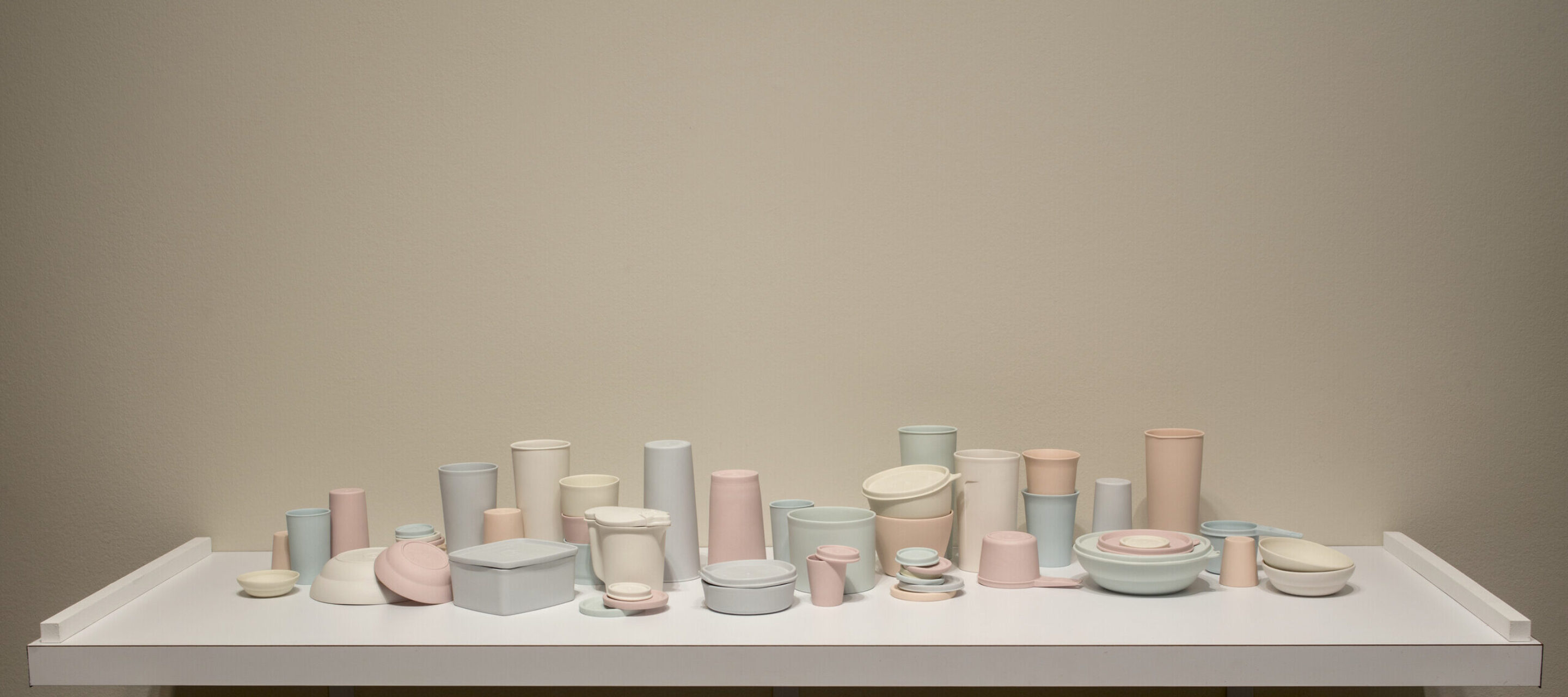 A collection of pastel-colored porcelain cups, bowls, containers, and lids that resemble plastic food containers. Some are stacked within and on top of each other. Some containers have lids firmly shut while on others the lids teeter off the edge of container rims.
