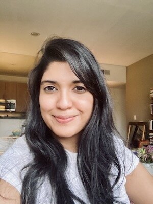 A light-skinned woman with straight black hair smiles with closed lips at the camera. She appears to be in an apartment. She wears a white T-shirt.