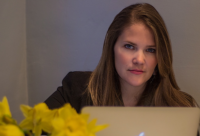 A light-skinned woman with straight light-brown hair stares seriously at the camera. She wears a black blazer and is seemingly sitting behind a laptop screen.