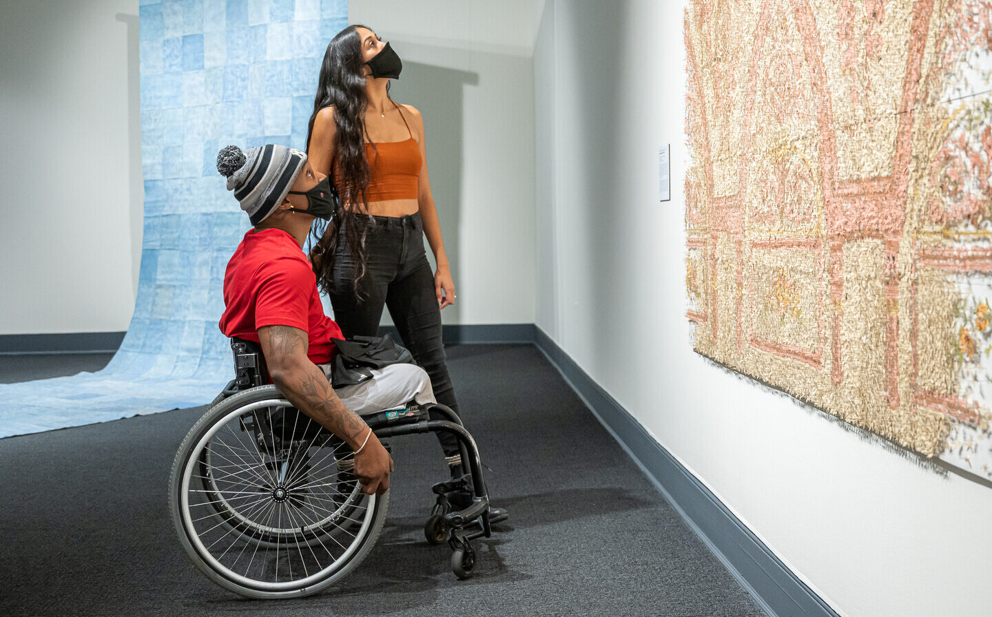 Two people look up at a large peach, tan, and ivory art installation in a gallery. The person with medium skin tone and tattoos on their right arm sits in a wheelchair and wears a red t-shirt, gray pants, gray-and-black striped winter hat, and black face mask. A person with medium skin tone, long curly black hair stands behind, wearing a burnt orange tank top, black jeans, and a black face mask. A large checkered blue artwork unfolds onto the gallery floor behind the pair.