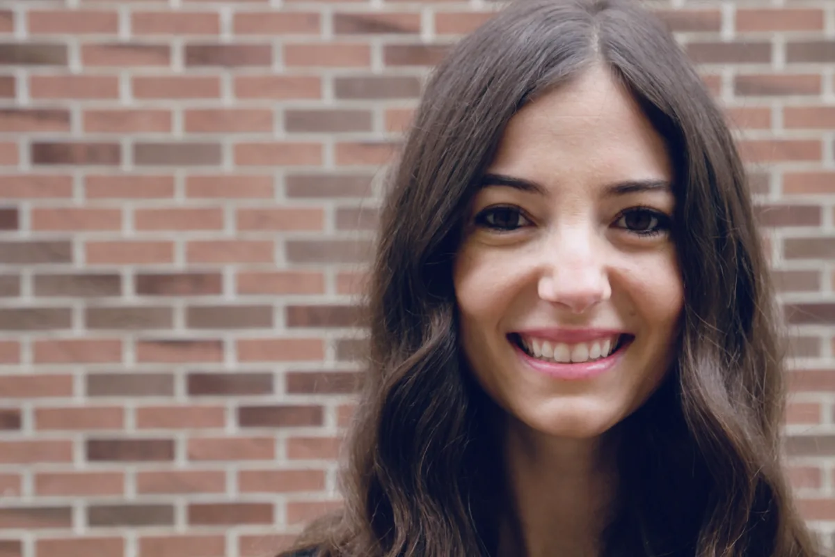 A light-skinned woman is pictured from the neck up, smiling happily in front of a red brick wall. Her brown hair is worn down, styled in subtle waves.