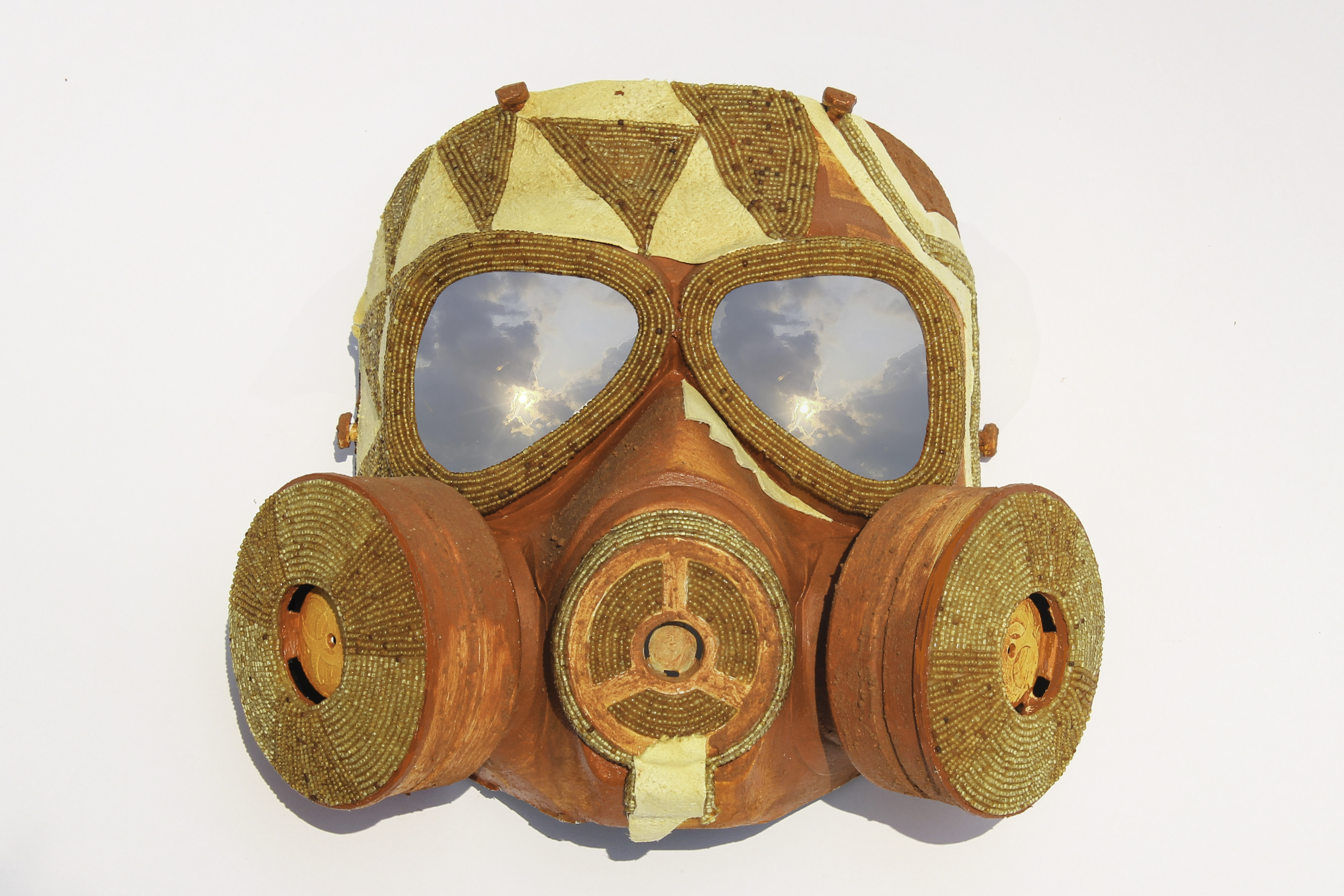 A gas mask decorated with shiny seed beads, animal hide, paint, and Oklahoma red dirt. The added materials form a geometric pattern on the forehead area, and a clouded sky can be seen reflected in the eye lenses of the mask.
