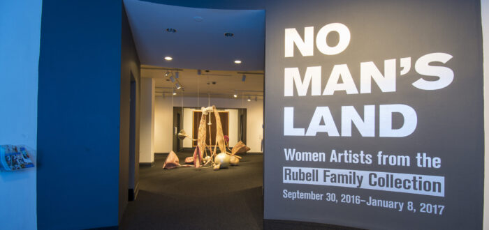 View of a gallery space. On a black wall, it says in white, bold letters: "NO MAN’S LAND: Women Artists from the Rubell Family Collection". There is a textile sculpture hanging from the ceiling in the room behind the wall.