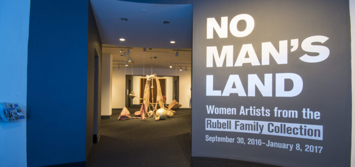 View of a gallery space. On a black wall, it says in white, bold letters: "NO MAN’S LAND: Women Artists from the Rubell Family Collection". There is a textile sculpture hanging from the ceiling in the room behind the wall.