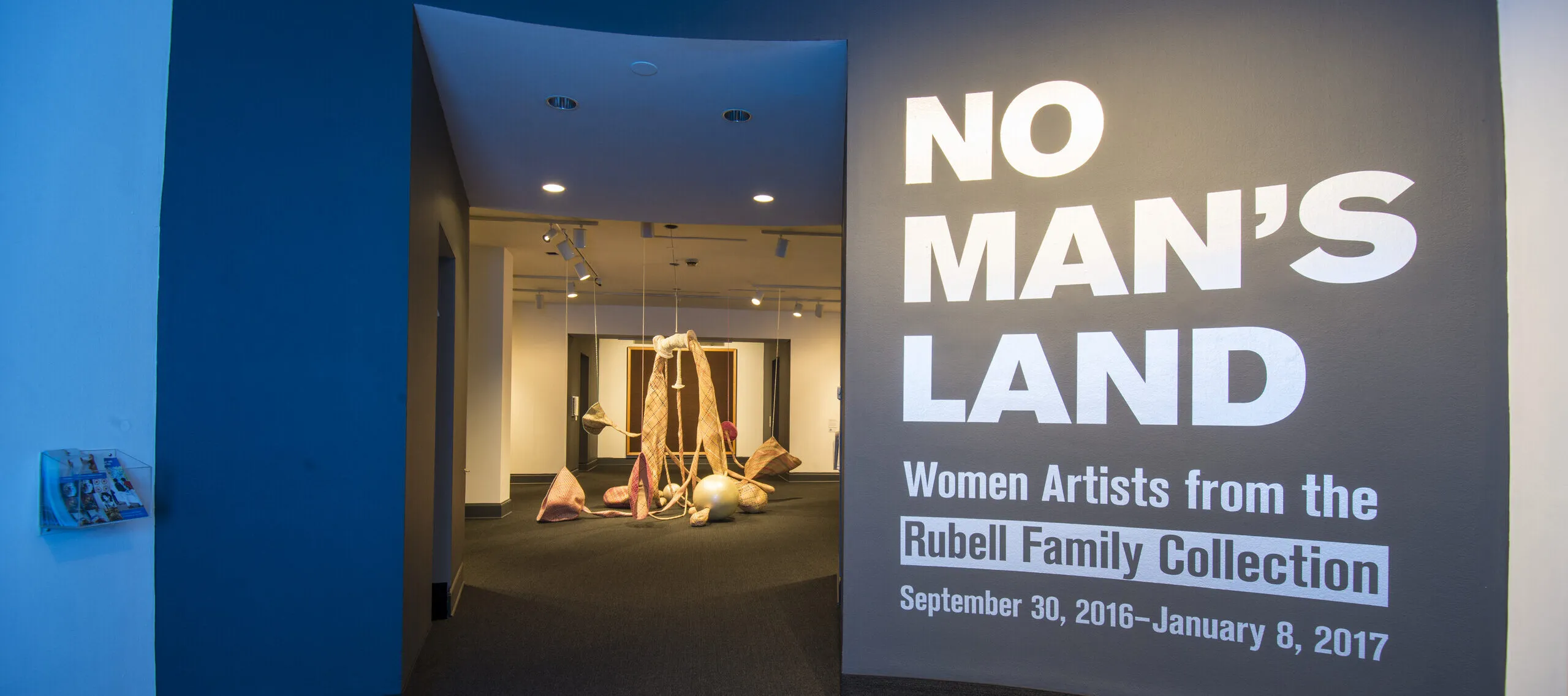 View of a gallery space. On a black wall, it says in white, bold letters: "NO MAN’S LAND: Women Artists from the Rubell Family Collection". There is a textile sculpture hanging from the ceiling in the room behind the wall.