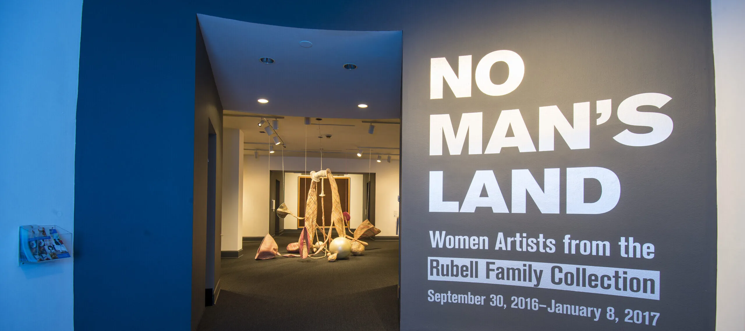 View of a gallery space. On a black wall, it says in white, bold letters: "NO MAN’S LAND: Women Artists from the Rubell Family Collection". There is a textile sculpture hanging from the ceiling in the room behind the wall.