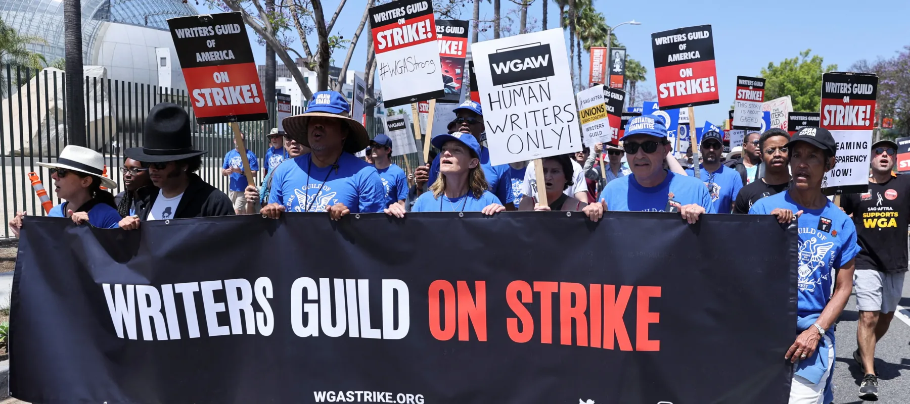 Six people stand wearing matching blue hats and shirts hold up and walk behind a large banner sign that says "Writers Guild on Strike." Behind them is a mass of other people holding picketing signs and chanting.