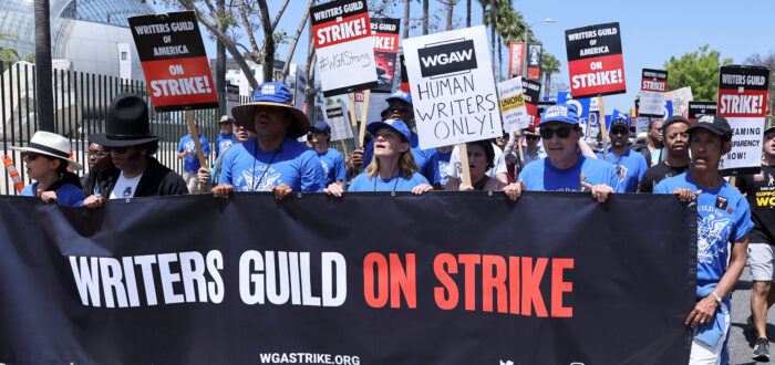 Six people stand wearing matching blue hats and shirts hold up and walk behind a large banner sign that says "Writers Guild on Strike." Behind them is a mass of other people holding picketing signs and chanting.