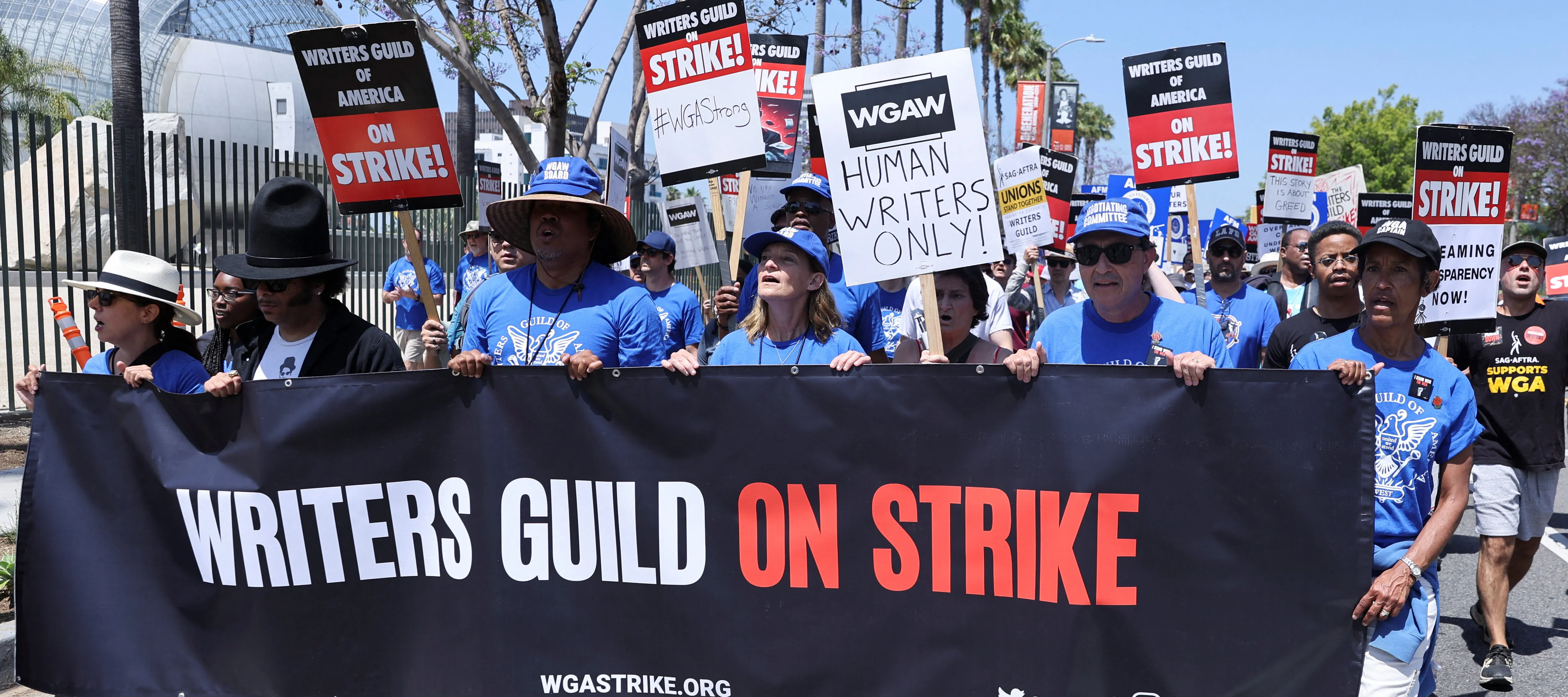 Six people stand wearing matching blue hats and shirts hold up and walk behind a large banner sign that says "Writers Guild on Strike." Behind them is a mass of other people holding picketing signs and chanting.