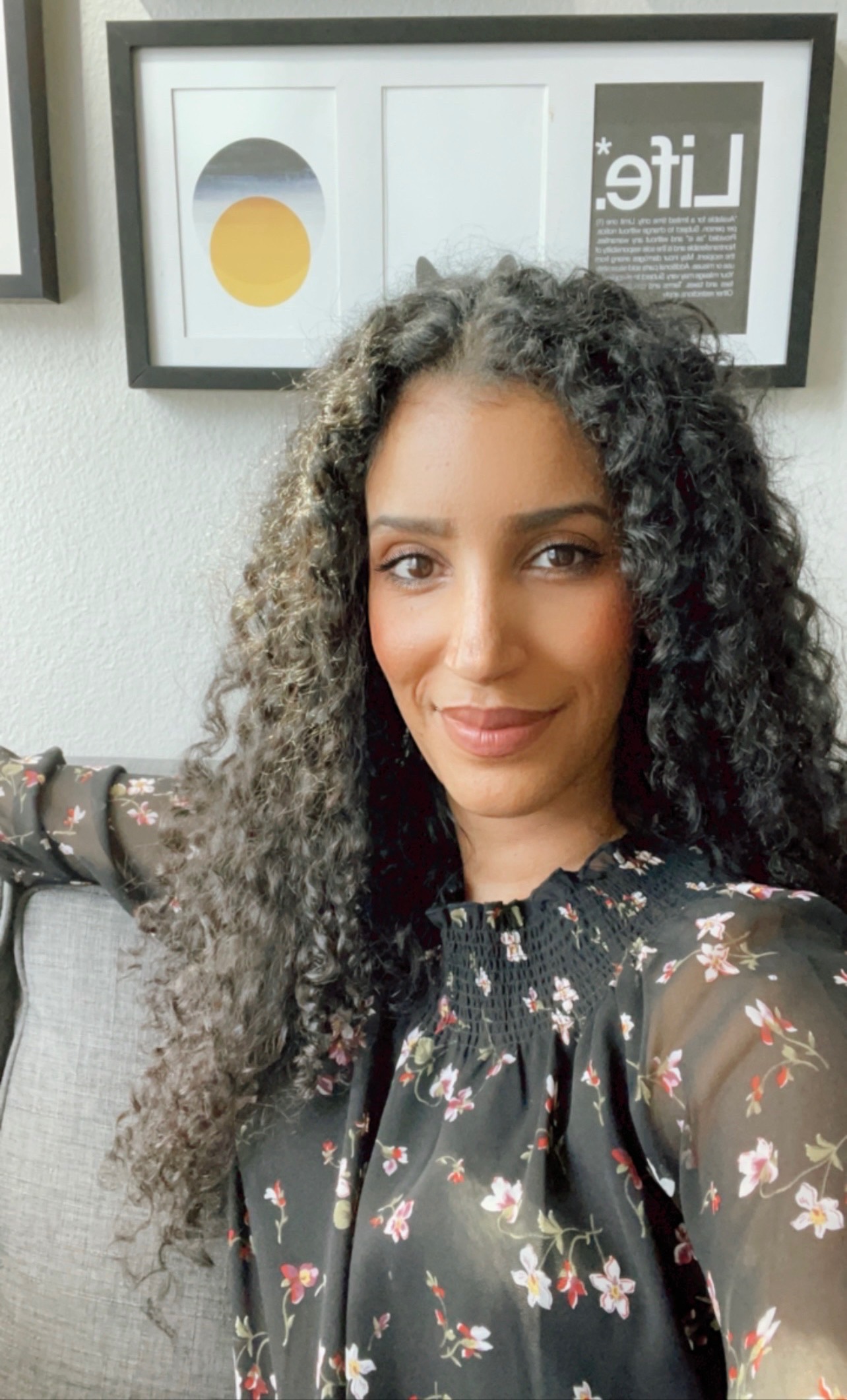 A medium-skinned woman with long, black curly hair takes a selfie while sitting on a couch. A framed artwork is visible behind her. She smiles warmly.