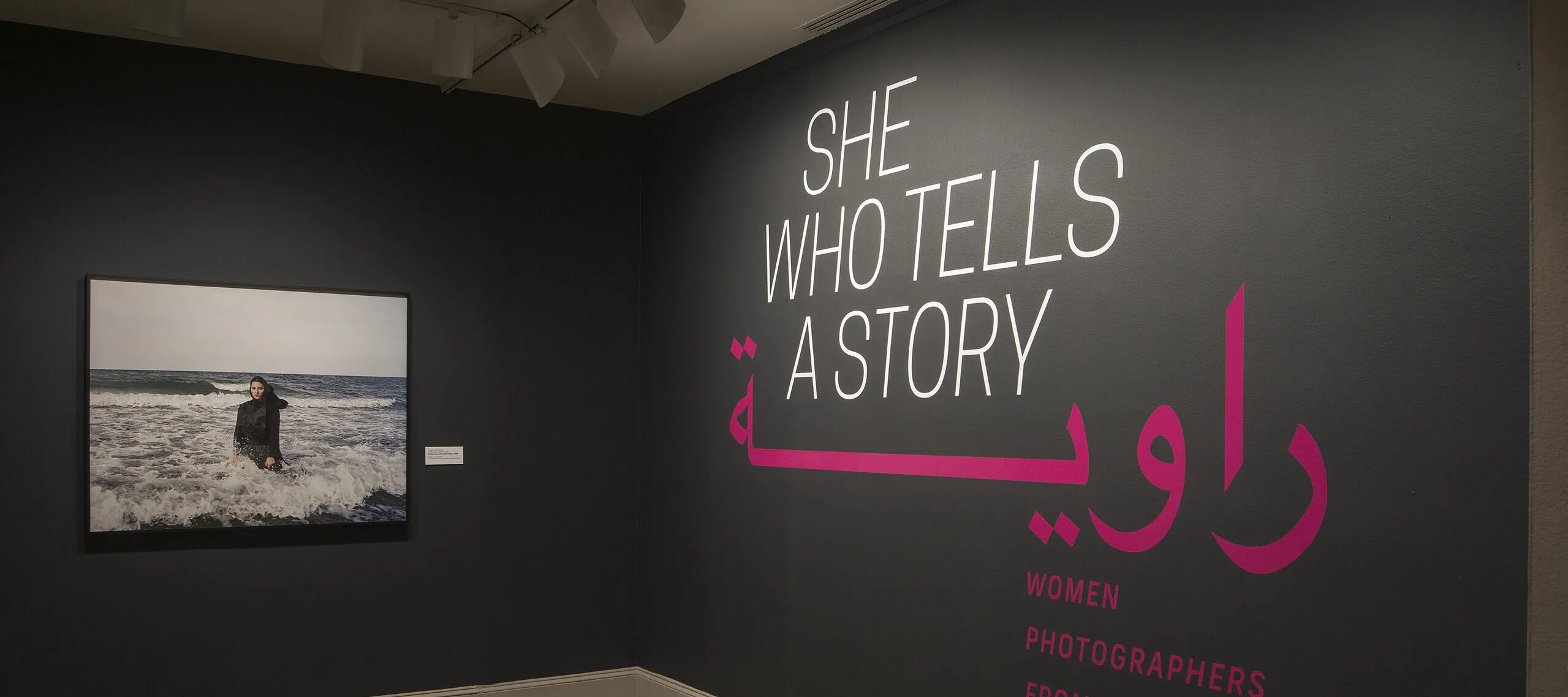 A gallery view of a black wall with a large photograph of a woman. The woman is wearing a long black dress and a head scarf. She is standing in the ocean, surrounded by waves. On the right wall is a text that says "She who tells a story".