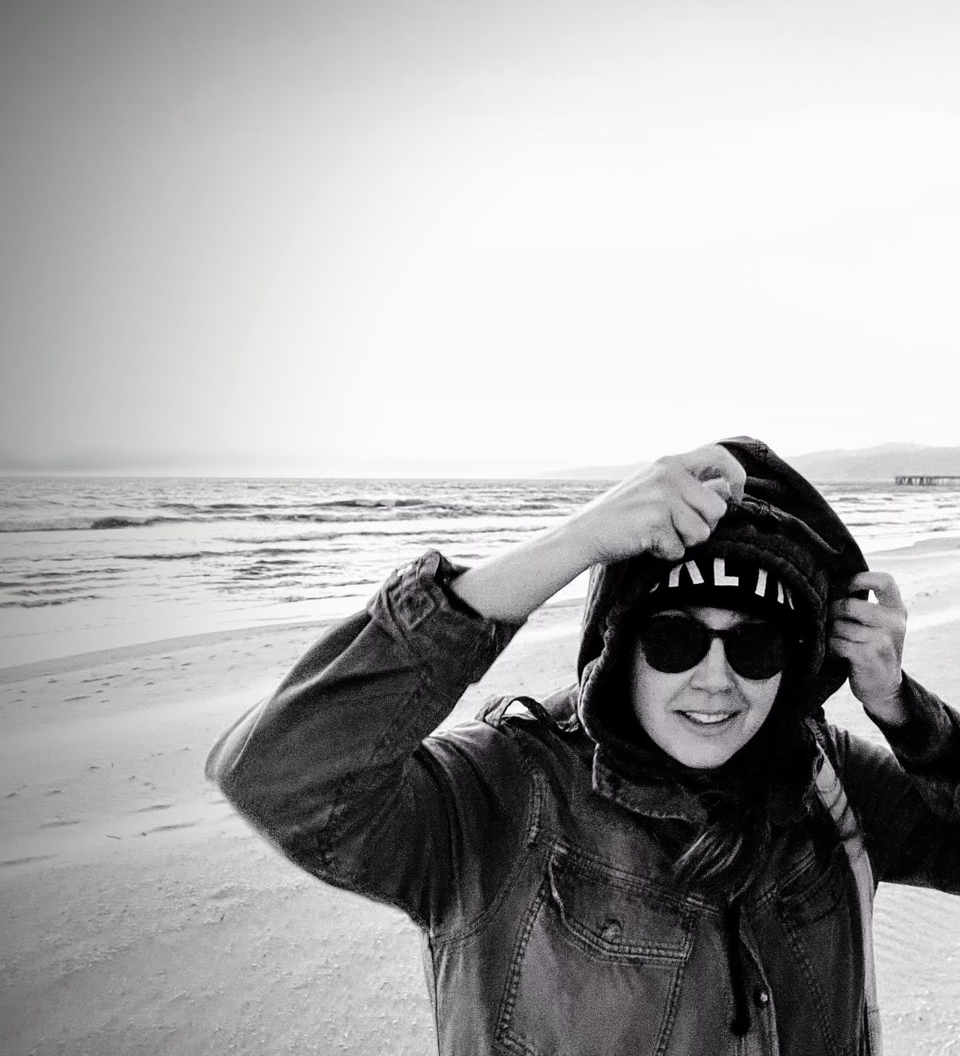 A black-and-white photo of a light-skinned woman standing on a beach, holding her jacket's hood up. She wears black sunglasses and smiles at the camera slightly.