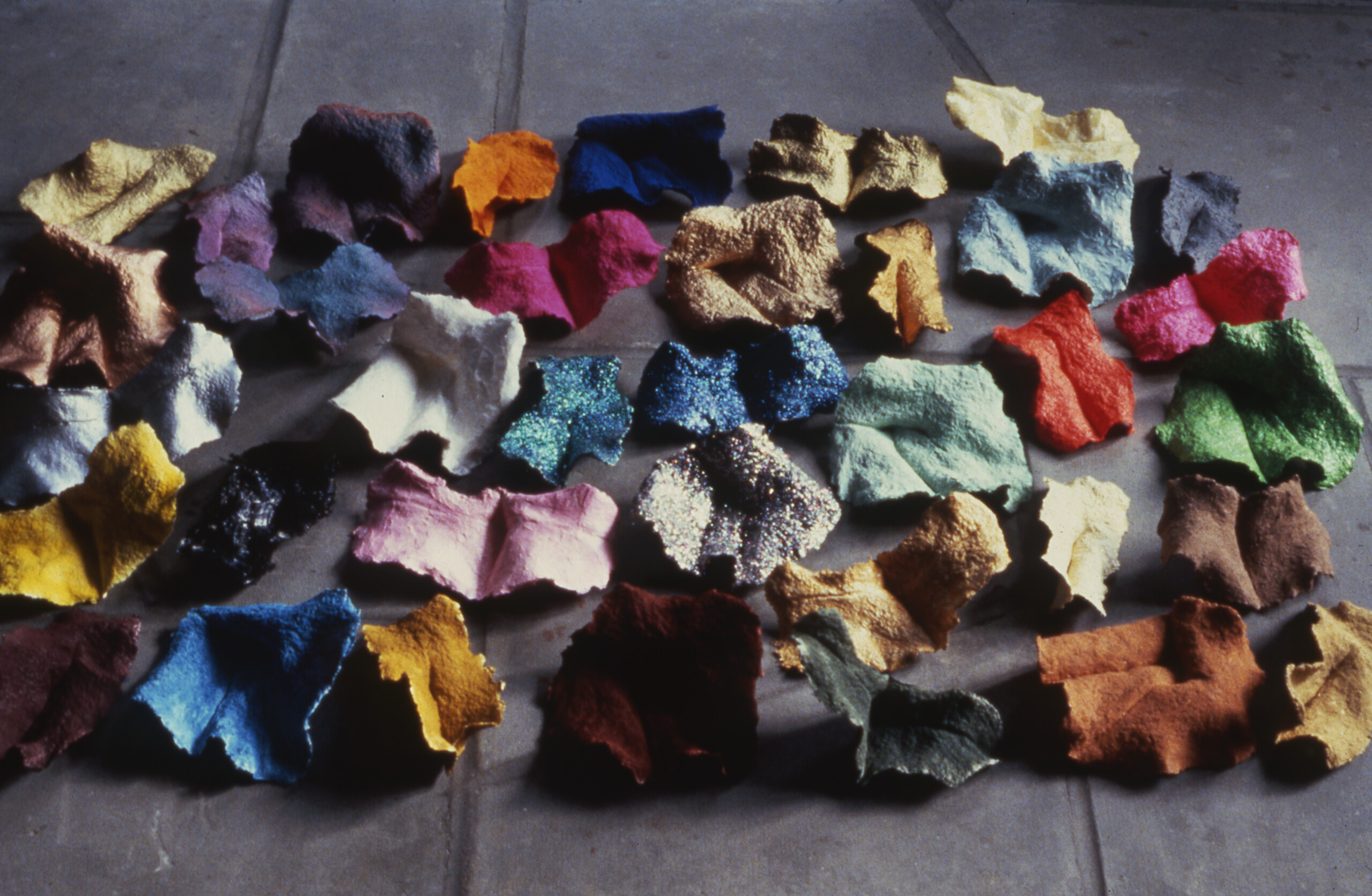 A cluster of homemade paper fragments in a variety of saturated colors sit on a tile floor. Some fragments are matte and some have glitter embedded in the paper.
