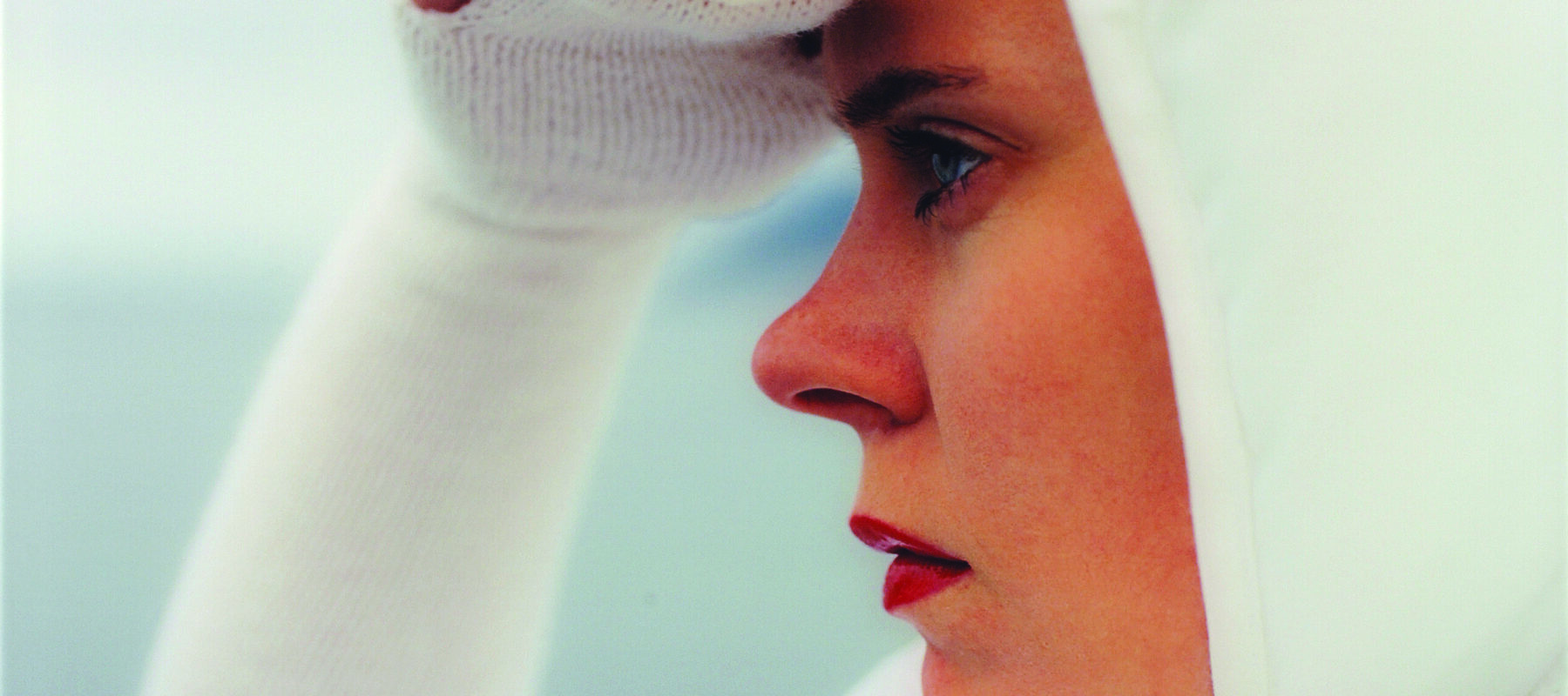 Square close-up of a light-skinned woman in profile looking to the left, seen from the shoulders up. She wears all white, including a bonnet and knit sleeves that become gloves covering her three middle fingers, and she holds her right hand up as if shielding her eyes from the sun.