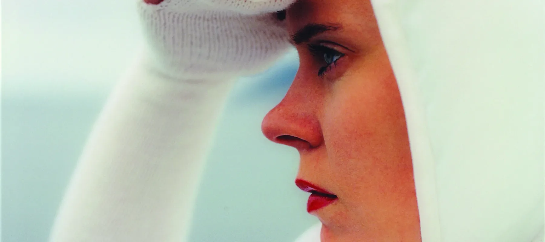 Square close-up of a light-skinned woman in profile looking to the left, seen from the shoulders up. She wears all white, including a bonnet and knit sleeves that become gloves covering her three middle fingers, and she holds her right hand up as if shielding her eyes from the sun.