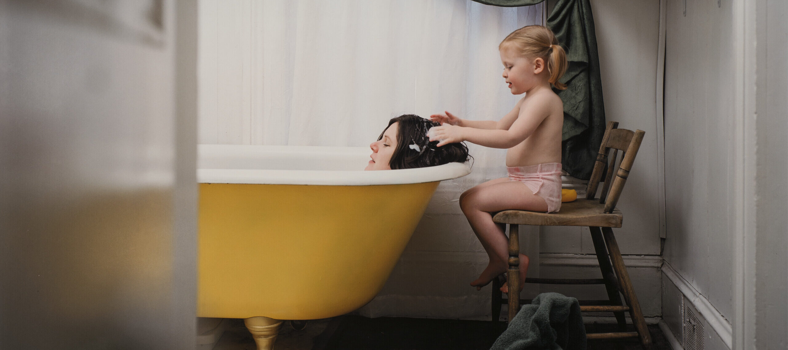 The head of a brunette woman with her eyes closed is visible above the rim of a yellow claw-footed bathtub. Behind the woman, a little girl in a pink diaper sits on a wooden chair and runs her soap-covered hands through the woman's hair.