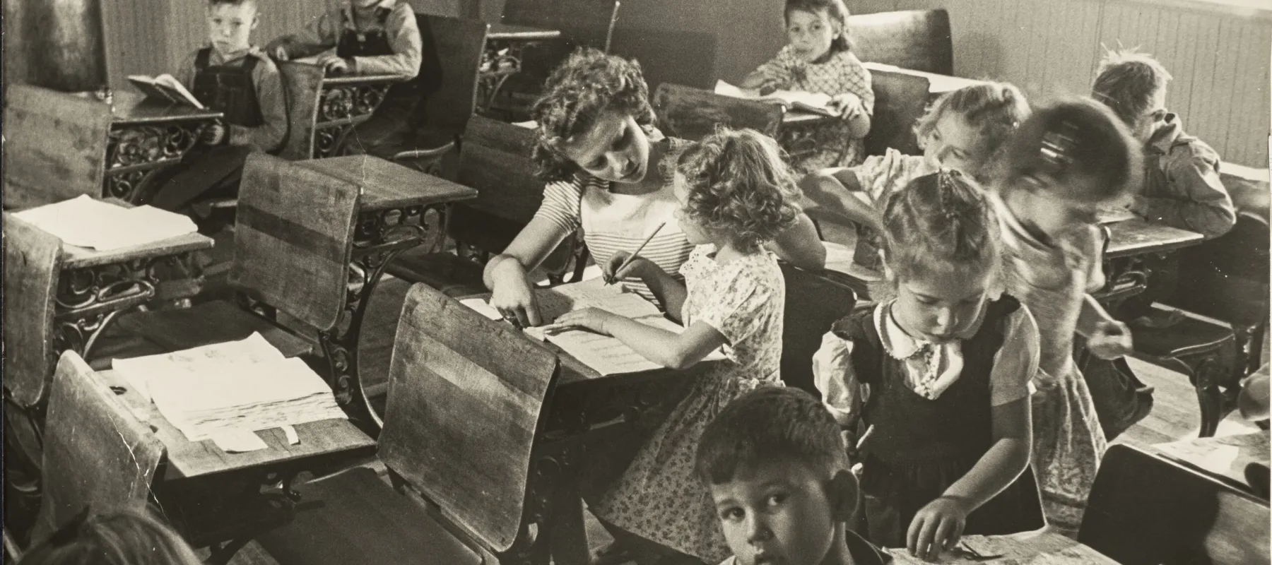 Black and white photograph of a primary school classroom in 1948. Light-skinned children sit at dark wood desks attending to books and papers or looking directly into the camera. At the center, the young teacher assists a girl with her lesson.