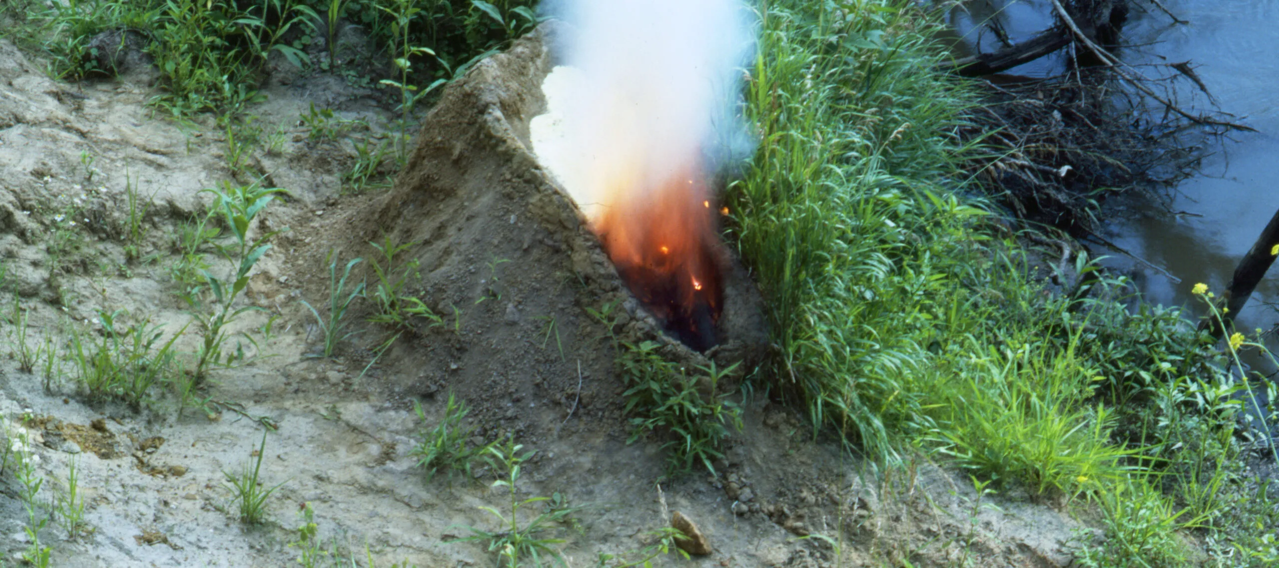 A mound of dirt in a grassy area by water. In the center of the mound is a human-shaped recess full of gunpowder that has been set aflame, exploding up in fire and white smoke, making the dirt mound resemble an erupting volcano.